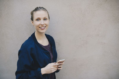 Portrait of happy female computer programmer holding smart phone while standing against beige wall in office