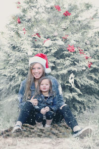 Portrait of smiling mother with cute daughter sitting by plants in park during snowfall
