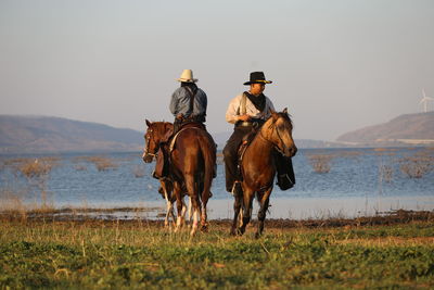 Men riding horses on grass by lake against sky