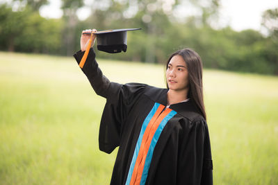 Young woman in graduation gown holding mortarboard on field 