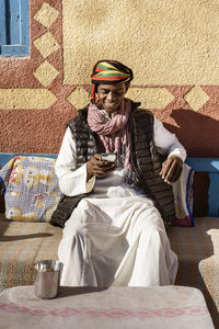 Cheerful egyptian male wearing traditional turban and clothes surfing internet on smartphone while sitting on sofa in courtyard near house on sunny day