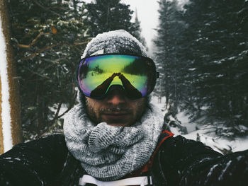 Portrait of man wearing ski goggles with reflection while standing against trees during winter