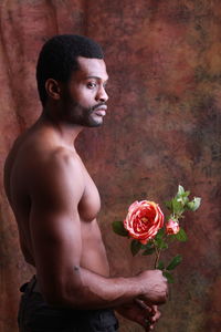 Shirtless young man holding flower while standing against wall