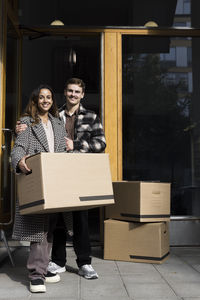 Portrait of smiling multiracial couple standing with boxes in front of apartment