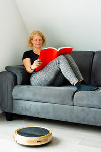 Woman reading book while sitting on sofa at home by vacuum cleaner