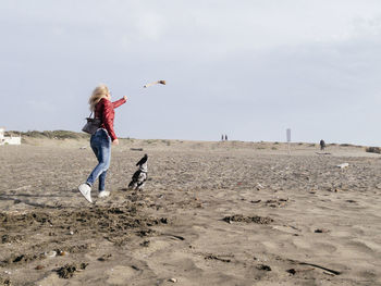 Full length of woman playing with dog on sand at beach against sky