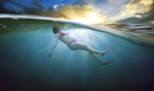 Pregnant woman floating undersea against cloudy sky on sunny day