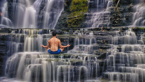 Rear view of shirtless man meditating while sitting against waterfall