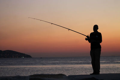 Silhouette man fishing on beach against sky during sunset