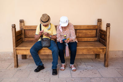 Full body of anonymous mature couple in headwear surfing cellphones while sitting on wooden bench on street of city