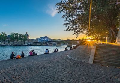 People relaxing on footpath by street against sky during sunset