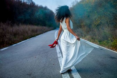 Side view of young woman in dress standing on road amidst trees