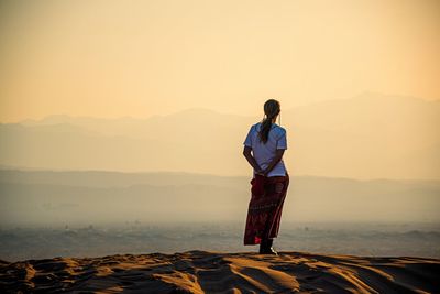 Rear view of man standing on beach against sky during sunset