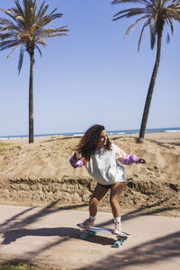 Full body of active female in casual clothes riding skateboard on road along sandy beach and tall palms during training