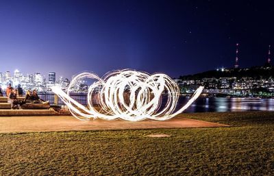 People by wire wool against lake in city at night