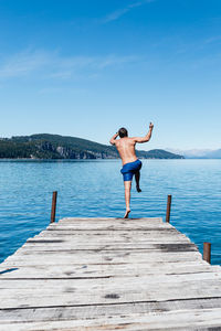 Rear view of woman jumping on pier over sea against sky