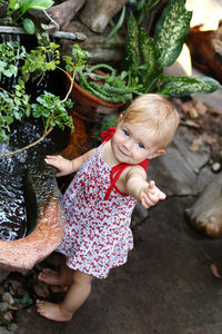 Little smiling girl with blond hair in sundress is standing in garden with pot plants and waterfall.