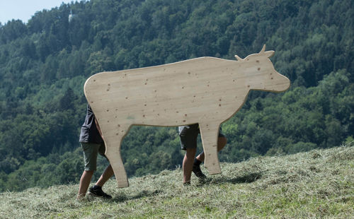 Two children carrying a wooden cow on a green meadow