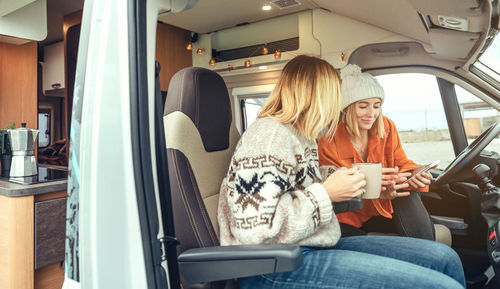 Women with a coffee looking mobile sitting in the front seat of a camper van