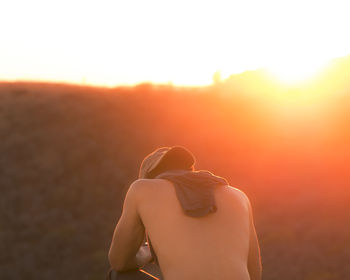 Rear view of shirtless man sitting against sky during sunset