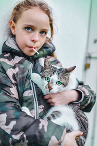 Portrait of cute girl eating lollipop while carrying cat at home