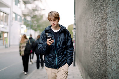 Teenage boy with blond hair using smart phone while walking on sidewalk against wall in city