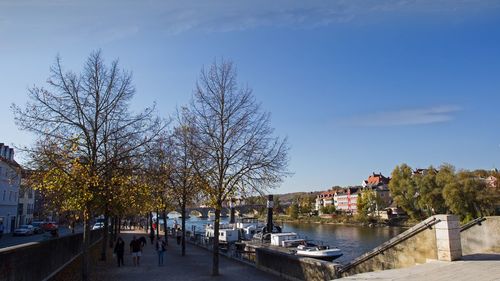 Bare trees by river in city against sky