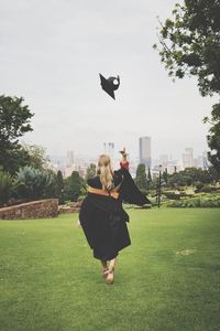 Rear view of young woman in graduation gown throwing mortarboard while walking on grassy field at park