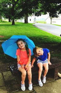 Full length portrait of sisters sitting with blue umbrella on railing at park