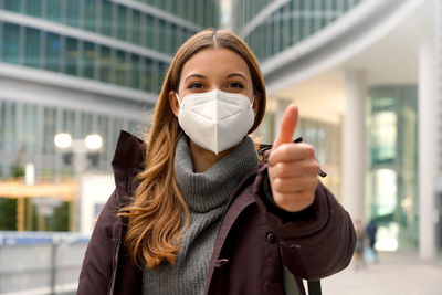 Woman wearing protective mask kn95 ffp2 showing thumb up and looking at camera in modern city