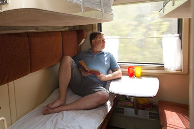 An adult blond man of 40-45 years old is lying down in his train compartment reading a book.