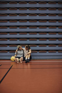 Full length of girl sitting with arm around female friend at sports court in school