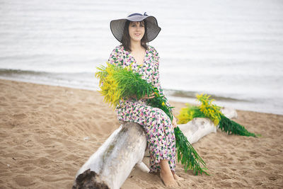 Young woman in summer dress and sun hat sitting on the beach and holding  bouquet of wild flower