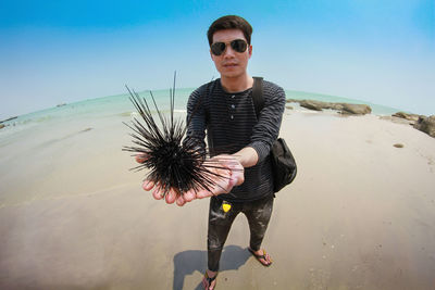 Portrait of young man holding urchin at beach against sky