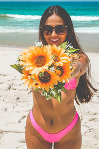 Portrait of young woman in bikini holding flowers while standing on beach