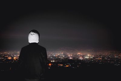 Rear view of man standing by illuminated buildings against sky at night