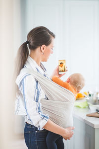 Side view of female blogger photo messaging while carrying daughter in kitchen at home