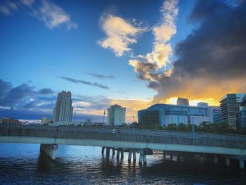 Bridge over river by buildings against sky during sunset