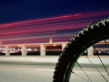 Close-up of bicycle tire by illuminated light trails on bridge at night
