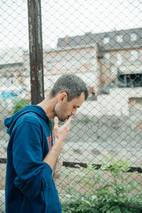 Side view of young man looking through chainlink fence