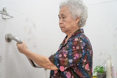 Senior woman holding handrail while standing by tiled floor in bathroom