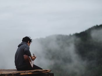 Side view of man playing guitar sitting at cliff