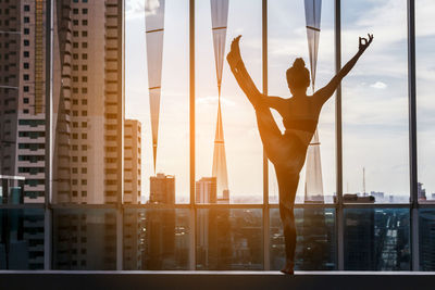 Rear view of young woman exercising against window in city during sunset