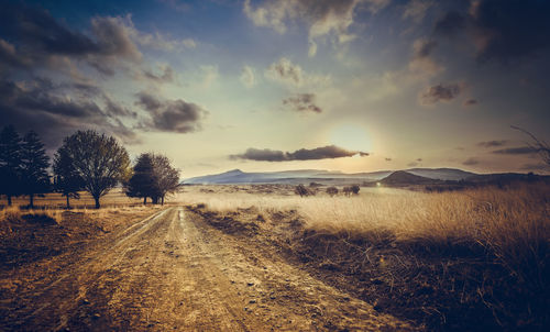 Dirt road amidst field against sky during sunset