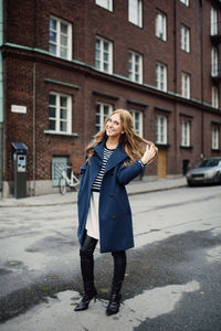 Full length portrait of smiling young woman standing with hand in hair on street against building in city