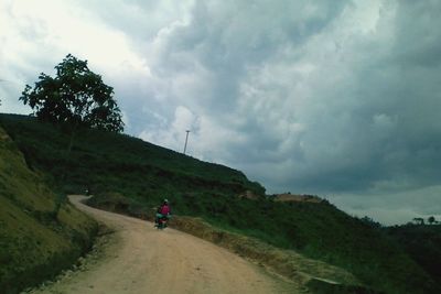 Rear view of woman walking on country road against cloudy sky