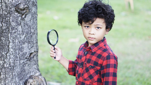 Portrait of boy holding magnifying glass while standing by tree trunk