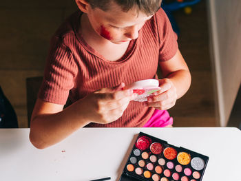 From above of crop child with makeup on face looking in mirror at table with eyeshadow palette at home