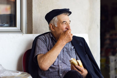 Senior man eating burger while sitting against wall