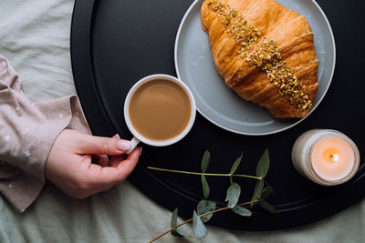 Flat lay of woman's hand holding cup cappuccino, croissant and scented candle with eucalyptus branch